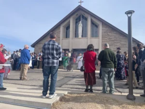 A congregation gathered outside a modern stone church under a blue sky.